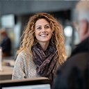 A woman with curly hair and a scarf smiles while speaking to someone across a counter in an indoor setting.