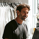 A man with curly hair and a beard, wearing a black t-shirt and necklace, stands in front of a rack of clothes, smiling at a computer screen.